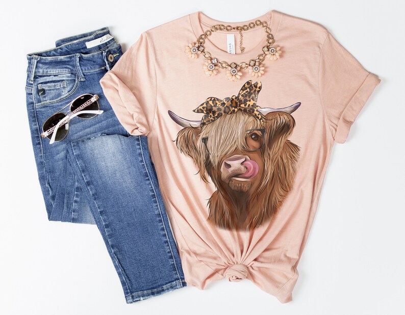 Puede incluir: Una camiseta rosa con una vaca Highland marr&oacute;n y blanca que lleva un pa&ntilde;uelo de leopardo y gafas. La vaca est&aacute; sacando la lengua.