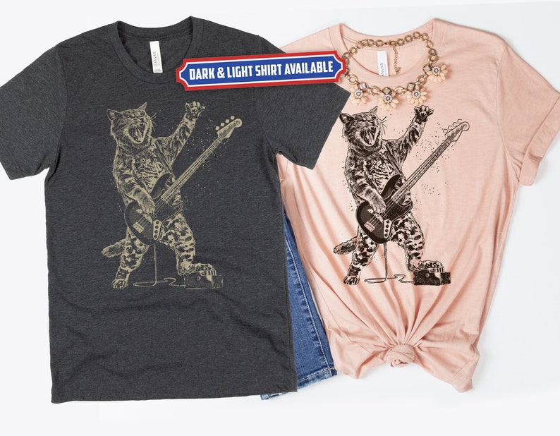 Może przedstawiać: Dwie koszulki z czarno-białym rysunkiem kota grającego na basie. Tekst "DARK & LIGHT SHIRT AVAILABLE" znajduje się na niebieskim banerze nad obrazkiem.