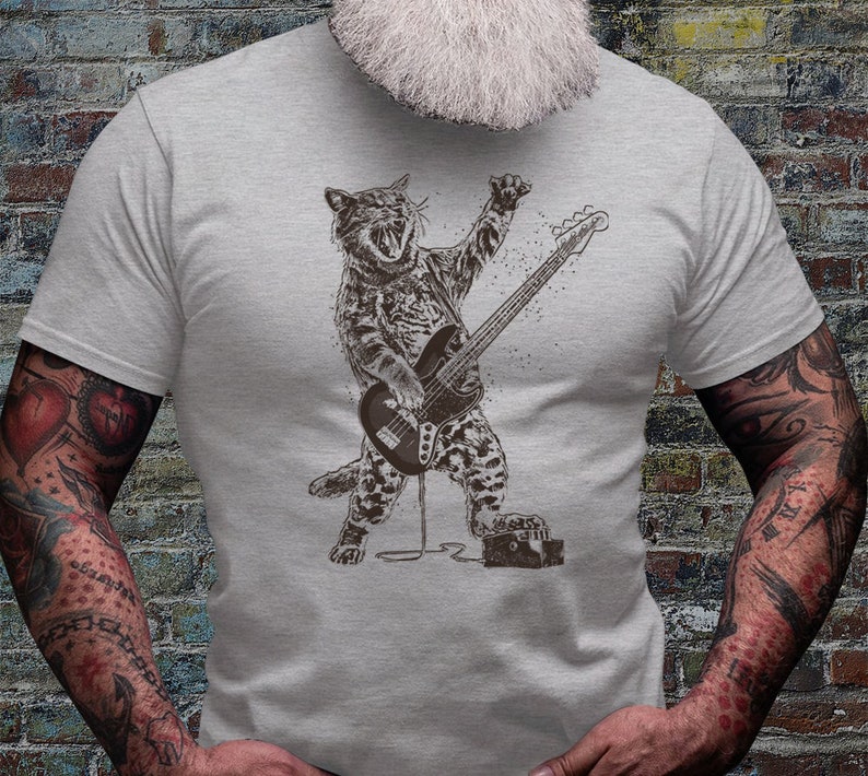 Może przedstawiać: Szary t-shirt z grafiką kota grającego na basie. Kot ma na sobie wz&oacute;r w panterkę i mikrofon w pysku.