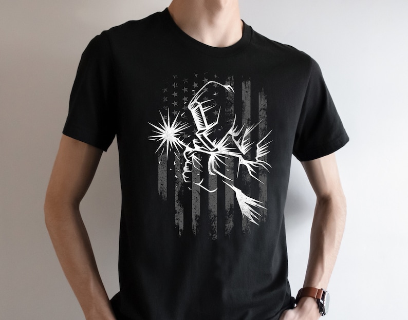 May include: Black t-shirt with a white graphic of a welder wearing a welding mask and holding a welding torch in front of a distressed American flag.