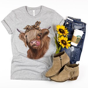 Puede incluir: Una camiseta gris con una vaca Highland marr&oacute;n y blanca que lleva gafas y una diadema de estampado de leopardo. La vaca est&aacute; sacando la lengua. La camiseta est&aacute; sobre una superficie blanca con un par de botines marrones de tobillo, unos vaqueros azules y una taza de caf&eacute;.