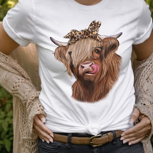 Puede incluir: Una camiseta blanca con una vaca Highland marr&oacute;n y blanca que lleva un pa&ntilde;uelo de leopardo y gafas. La vaca est&aacute; lamiendo sus labios.