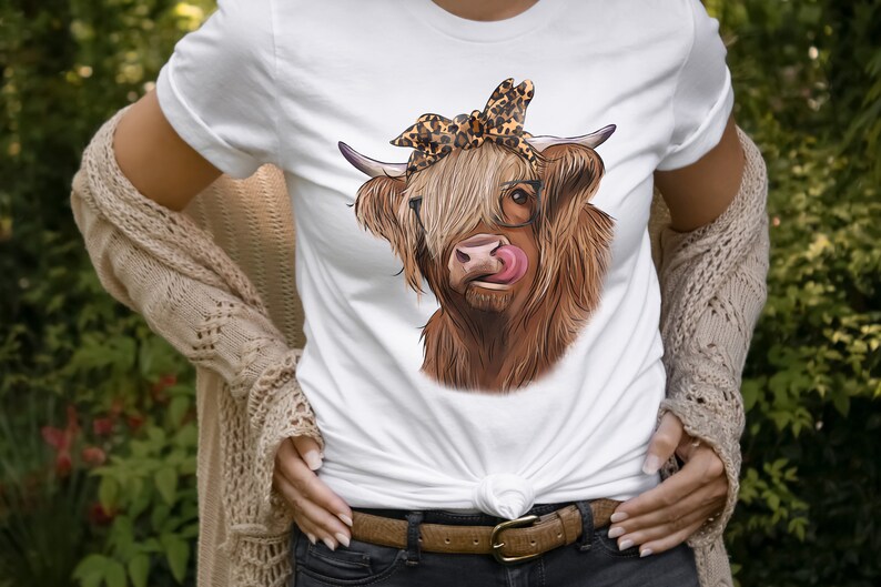 Puede incluir: Una camiseta blanca con una vaca Highland marr&oacute;n y blanca que lleva un pa&ntilde;uelo de leopardo y gafas. La vaca est&aacute; lamiendo sus labios.