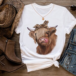 Puede incluir: Camiseta blanca con una vaca Highland marr&oacute;n y blanca que lleva un pa&ntilde;uelo de leopardo y gafas. La vaca se est&aacute; lamiendo la nariz.
