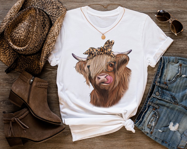 Puede incluir: Camiseta blanca con una vaca Highland marr&oacute;n y blanca que lleva un pa&ntilde;uelo de leopardo y gafas. La vaca se est&aacute; lamiendo la nariz.
