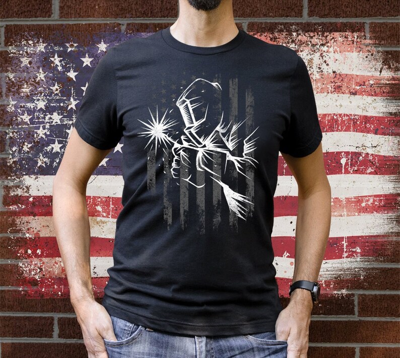 May include: A black t-shirt with a white graphic of a welder wearing a welding mask in front of a distressed American flag design.