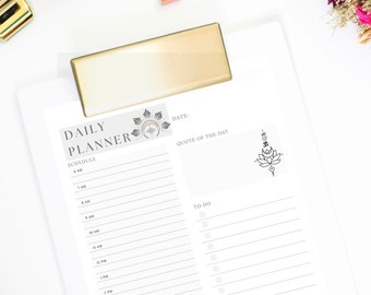 Op de afbeelding: Een witte dagelijkse planner met een grijs en zwart bloemenontwerp. De planner heeft een sectie voor planning, een sectie voor taken en een sectie voor een citaat van de dag. De planner ligt op een wit oppervlak met een gouden clip.