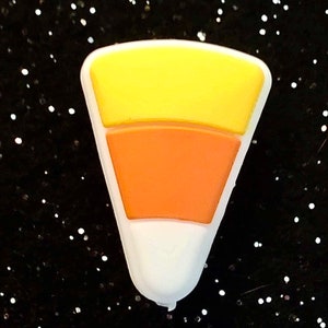 Puede incluir: Un dije de caramelo de maíz con un diseño amarillo, naranja y blanco. El dije tiene forma de caramelo de maíz y tiene un borde blanco. El fondo es negro con purpurina blanca.