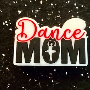 Puede incluir: Encanto blanco y negro con las palabras "Dance Mom" en letras rojas y negras. Una silueta de una bailarina está en el centro del encanto.