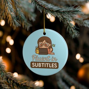 May include: A round, light blue Christmas ornament with a cartoon illustration of a person wearing glasses and working on a laptop. The ornament has the text "Fluent in SUBTITLES" in a brown font.