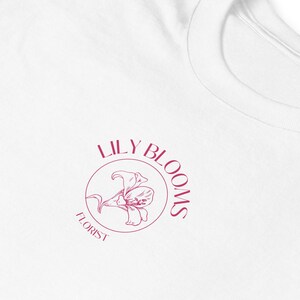 Puede incluir: Camiseta blanca con un logotipo rosa que dice "Lily Blooms Florist" con una flor de lirio estilizada dentro de un c&iacute;rculo.