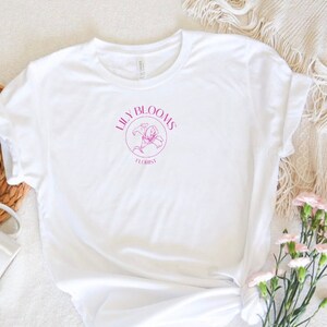 Puede incluir: Camiseta blanca con un gr&aacute;fico rosa de una flor de lirio y el texto "Lilly Blooms Florist".