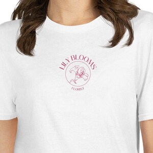 Puede incluir: Camiseta blanca con un logotipo rosa que dice "Lily Blooms Florist" y un gr&aacute;fico de flor de lis rosa.