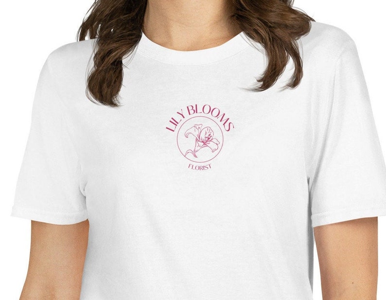 Puede incluir: Camiseta blanca con un logotipo rosa que dice "Lily Blooms Florist" y un gr&aacute;fico de flor de lis rosa.