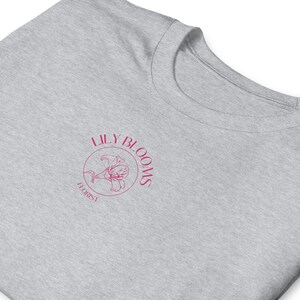 Puede incluir: Una camiseta gris con un logotipo circular rosa que dice "Lily Blooms Florist" y presenta una flor de lirio estilizada.
