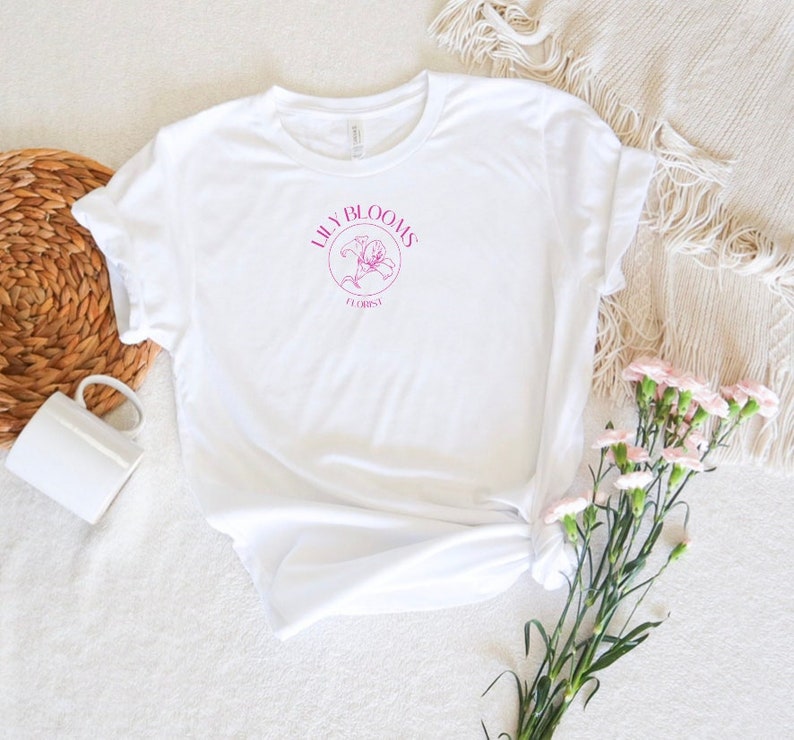Puede incluir: Camiseta blanca con un gr&aacute;fico rosa de una flor de lirio y el texto "Lilly Blooms Florist".