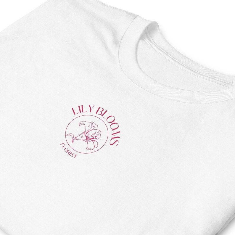 Puede incluir: Camiseta blanca con un logotipo rosa que dice "Lily Blooms Florist" con una flor de lirio estilizada dentro de un c&iacute;rculo.