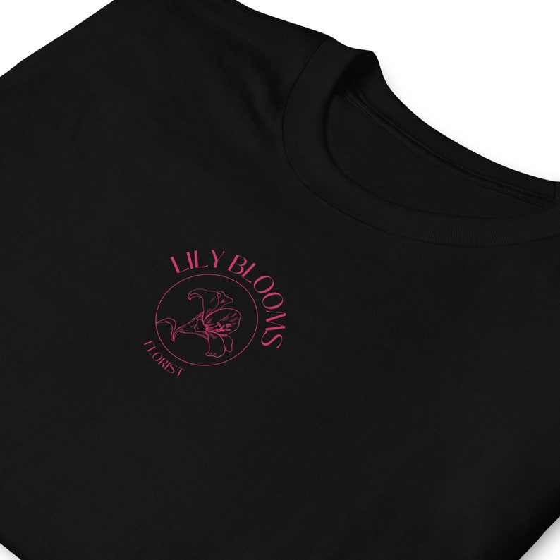 Puede incluir: Camiseta negra con un logotipo circular rosa que dice "Lily Blooms Florist" y presenta una flor de lirio estilizada.