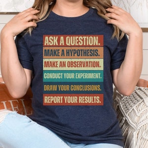 May include: A heather navy blue t-shirt with a vintage-style graphic that reads "Ask a question. Make a hypothesis. Make an observation. Conduct your experiment. Draw your conclusions. Report your results."