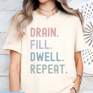 May include: A cream colored t-shirt with the text "DRAIN. FILL. DWELL. REPEAT." in a vintage style font. The text is in shades of pink, purple, blue, and green.