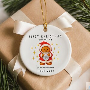 May include: A white ceramic Christmas ornament with a gold string, featuring a gingerbread person in a Santa suit. Text reads "FIRST CHRISTMAS without my swimmers JOHN 2025". The ornament is on a wrapped gift.