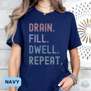 May include: A navy blue t-shirt with the text "DRAIN. FILL. DWELL. REPEAT." in a gradient of pink, blue, and green.