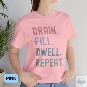 May include: Pink t-shirt with the text "DRAIN. FILL. DWELL. REPEAT." in a faded, layered font.