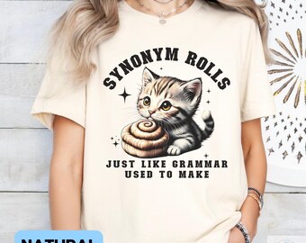 May include: A beige t-shirt with the text "Synonym Rolls Just Like Grammar Used To Make" and a cartoon illustration of a cat holding a cinnamon roll.