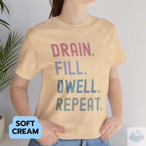 May include: A soft cream colored t-shirt with the text "DRAIN. FILL. DWELL. REPEAT." in a vintage style font. The text is in shades of pink, blue, and green.