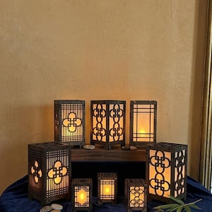 May include: A collection of dark brown wooden lanterns with intricate cut-out designs. The lanterns vary in size and shape, with warm, glowing light emanating from within. Some lanterns feature a floral-like pattern. The lanterns are arranged on a dark blue surface.