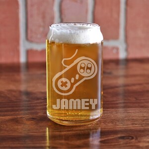 May include: A clear glass beer can with a white frosted design of a video game controller and the name "Jamey". The can is filled with a light golden beer and has a white head.