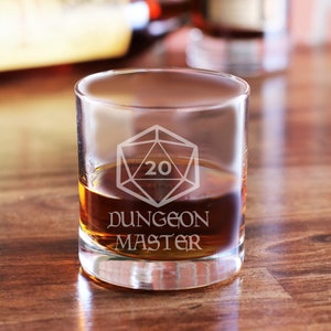 May include: A clear glass tumbler with a brown liquid inside. The glass is etched with a geometric design featuring the number 20 and the words "DUNGEON MASTER". The glass is sitting on a wooden surface.