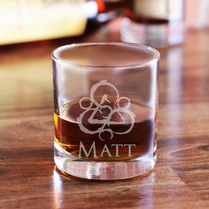 May include: A clear glass rocks glass with a brown liquid inside. The glass has an etched design of a circle with a triangle inside and the name "MATT" below it.
