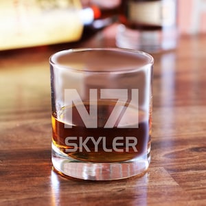 May include: A clear glass tumbler with a brown liquid inside. The glass has the text "NZ1 SKYLER" etched on the side.