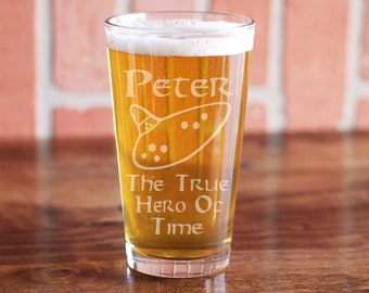 May include: A clear glass beer stein with a personalized inscription that reads "Peter The True Hero Of Time". The inscription is etched into the glass and includes a drawing of a ocarina.