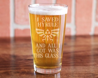 May include: A clear glass pint with a white inscription that reads "I saved Hyrule and all I got was this glass..." with a stylized image of the Triforce symbol. The glass is filled with a golden-colored beverage.