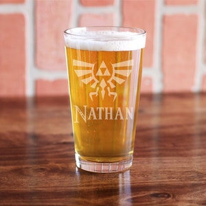 May include: A clear pint glass filled with beer and foam, featuring an etched design of a crest and the name "NATHAN". The glass is set on a wooden surface against a brick wall background.