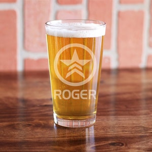 May include: A glass of light yellow beer with a white head in a clear pint glass. The glass has a white star logo and the word "ROGER" etched on the side.