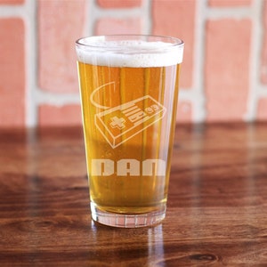 May include: A tall glass of beer with a white foam head. The glass is etched with a video game controller and the name "DAN".