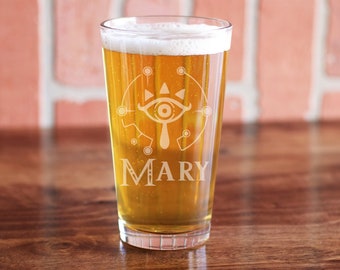 May include: A clear glass pint glass with a design of a stylized eye with a teardrop and the word "MARY" etched on the bottom. The glass is filled with a light amber colored beverage.