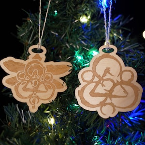 May include: Two wooden Christmas ornaments hanging on a decorated tree. The ornaments are light brown, with a decorative design etched into the wood. One is shaped like a stylized angel, the other is a rounded shape. Both have a hole at the top for hanging.