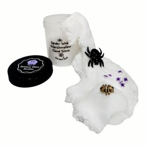 May include: A clear plastic container labeled Spider Web Marshmallow Cloud Slime with a black lid featuring the Sensory Slime Studios logo. Beside it, white cloud slime is spread out, decorated with a black spider, a gold spider, and small purple spider charms.