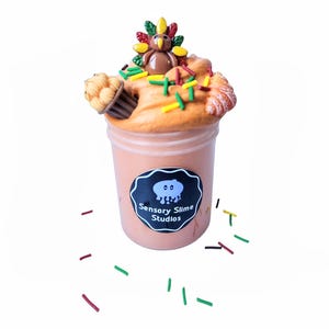 May include: A clear plastic container filled with orange-colored slime, topped with a decorative arrangement featuring a turkey figurine, a cupcake, and colorful sprinkles. The container has a black label with the text "Sensory Slime Studios".