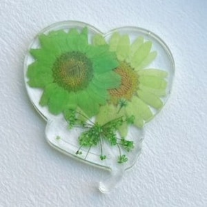 Pode incluir: Um ornamento de resina em forma de cora&ccedil;&atilde;o com flores e folhas verdes prensadas. As flores est&atilde;o dispostas em um cacho no centro do ornamento.