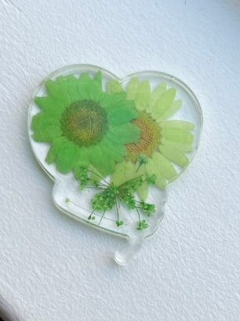 Pode incluir: Um ornamento de resina em forma de cora&ccedil;&atilde;o com flores e folhas verdes prensadas. As flores est&atilde;o dispostas em um cacho no centro do ornamento.