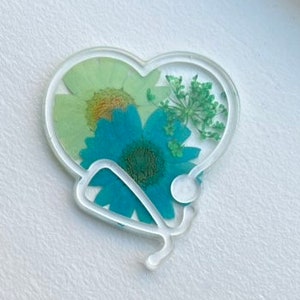 Pode incluir: Um ornamento de resina em forma de cora&ccedil;&atilde;o com um design de estetosc&oacute;pio e flores prensadas dentro. As flores s&atilde;o verdes, azuis e amarelas.