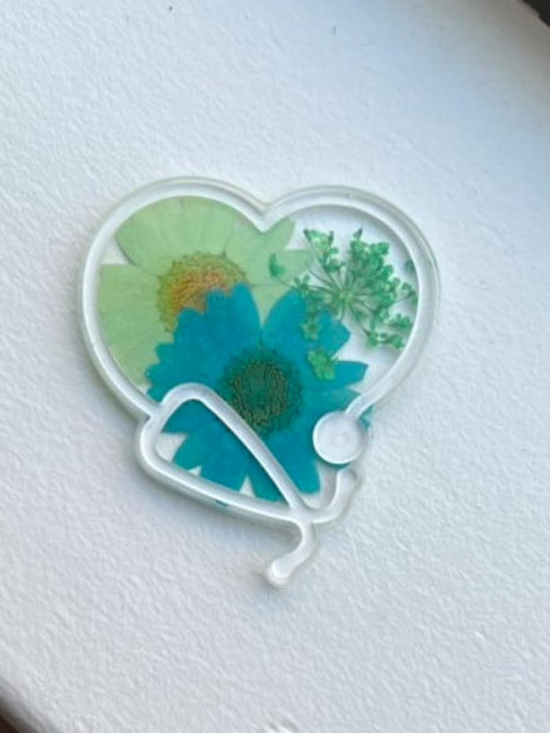 Pode incluir: Um ornamento de resina em forma de cora&ccedil;&atilde;o com um design de estetosc&oacute;pio e flores prensadas dentro. As flores s&atilde;o verdes, azuis e amarelas.