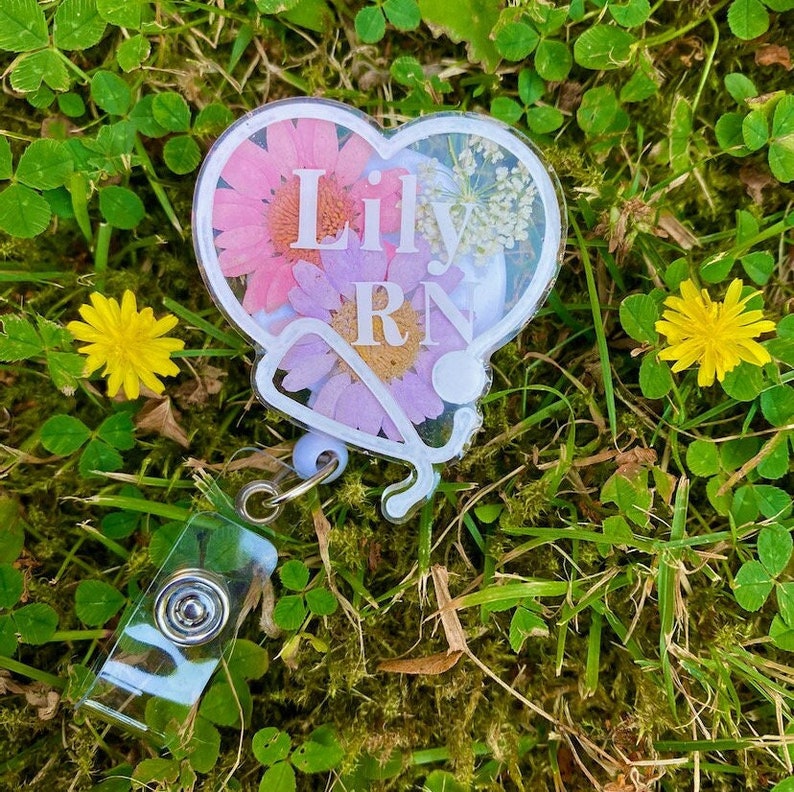 Pode incluir: Um porta-crach&aacute; branco em forma de cora&ccedil;&atilde;o com um design de estetosc&oacute;pio e flores prensadas. O porta-crach&aacute; tem o nome "Lily RN" escrito nele.