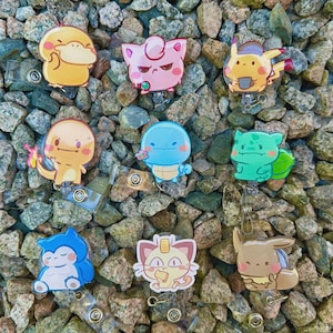 May include: A collection of colorful cartoon character badge reels. The badge reels feature various characters in yellow, pink, blue, green, and brown. The badge reels are set against a background of small, multicolored rocks.