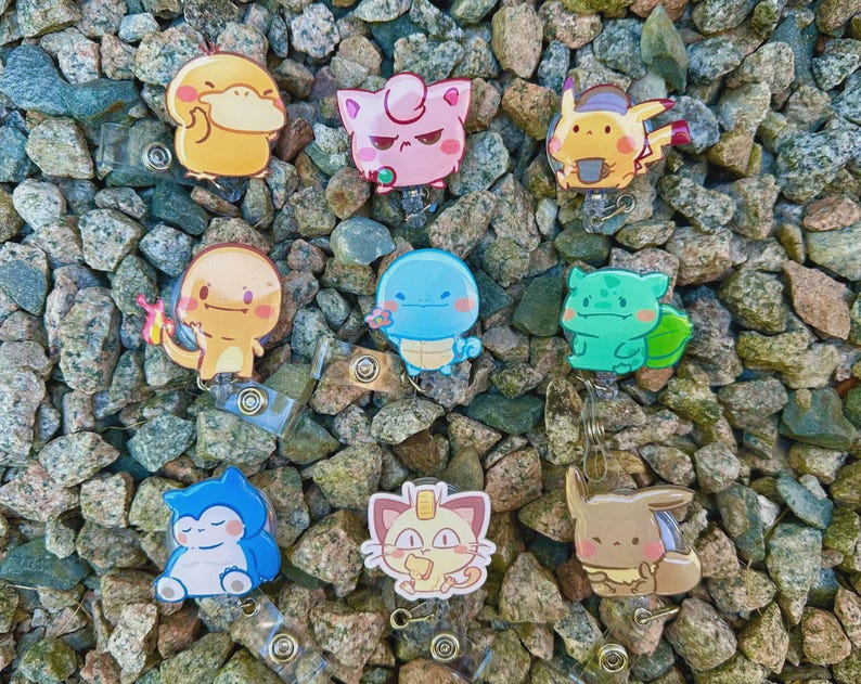 May include: A collection of colorful cartoon character badge reels. The badge reels feature various characters in yellow, pink, blue, green, and brown. The badge reels are set against a background of small, multicolored rocks.